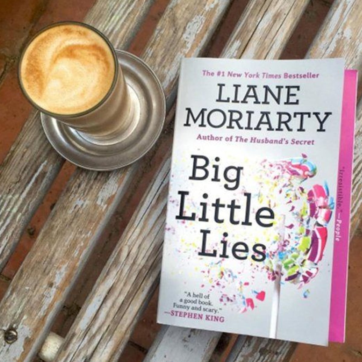 A glass of coffee sits on a saucer next to the book "Big Little Lies" by Liane Moriarty, which is resting on a weathered wooden bench. The book cover features bright, colorful confetti and a lollipop.