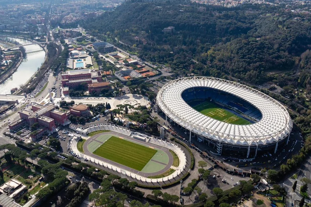 Aerial view of a large football stadium surrounded by roads, buildings, and trees, with a smaller sports field nearby and a river running alongside the cityscape in the background.