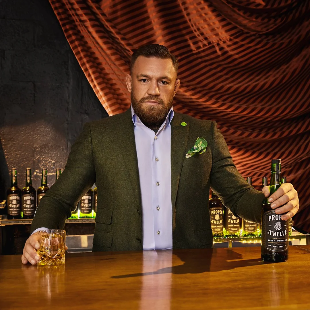 A man with a beard in a suit stands behind a bar, holding a bottle of Proper No. Twelve whiskey and a glass of whiskey, with several bottles displayed in the background.