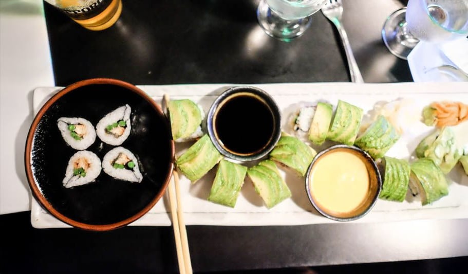 A plate of sushi rolls with avocado slices, a dish of soy sauce, a dish of yellow sauce, and four sushi pieces on a small black plate with chopsticks on the side. Drinks and utensils are in the background.
