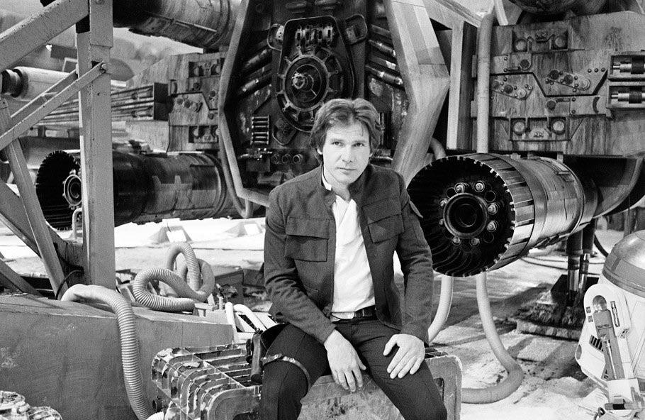 A man in a jacket and white shirt sits in front of a large spacecraft with detailed machinery, cables, and engines around him in a sci-fi setting.