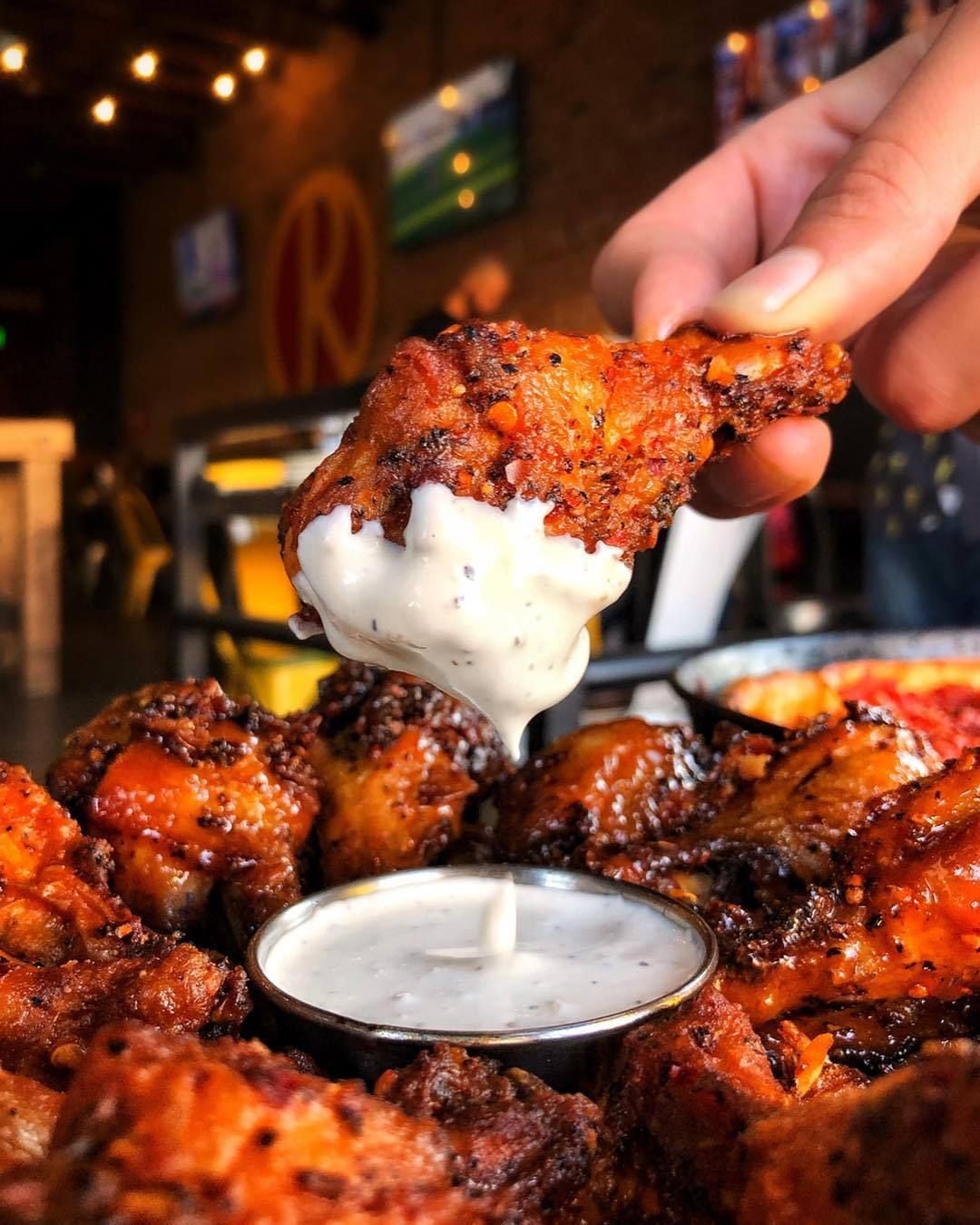 A close-up of a hand dipping a seasoned chicken wing into a cup of creamy white sauce, with a plate full of more wings in the background at a restaurant.