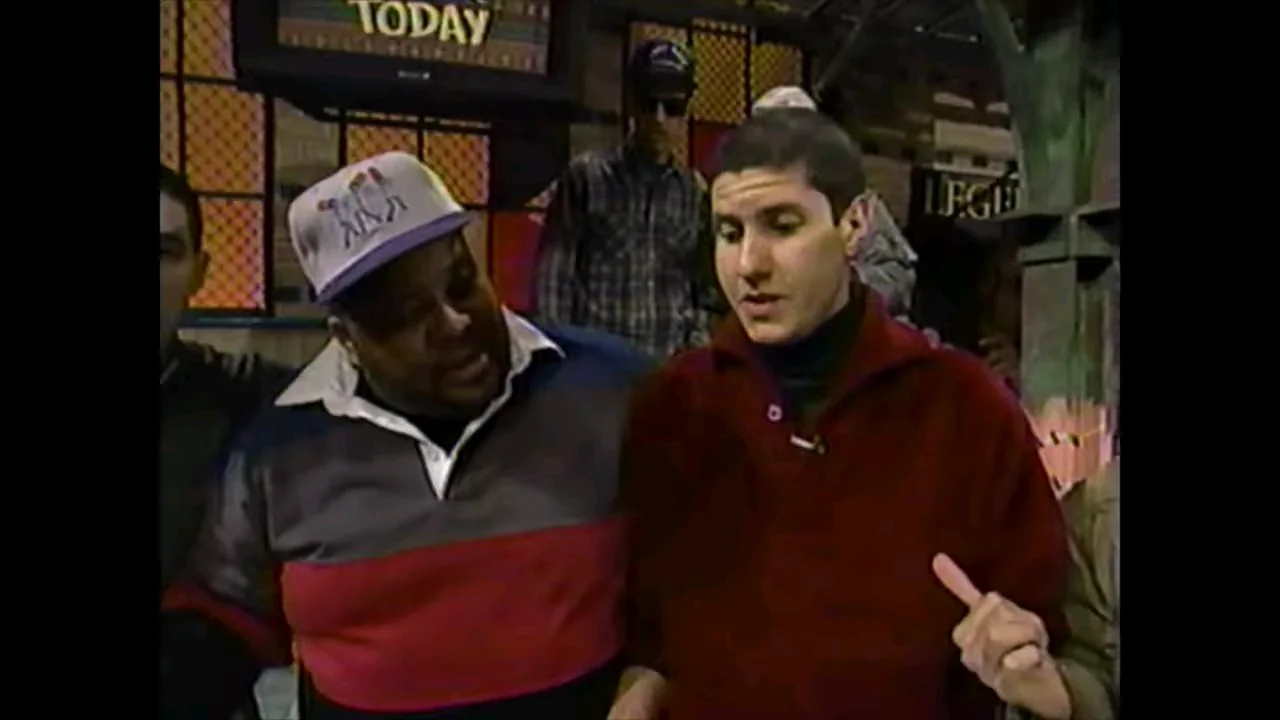 Two men stand close together indoors. The man on the left wears a striped shirt and light-colored cap, while the man on the right, in a red sweater, gestures with his hand. Several people and a "TODAY" sign are visible in the background.