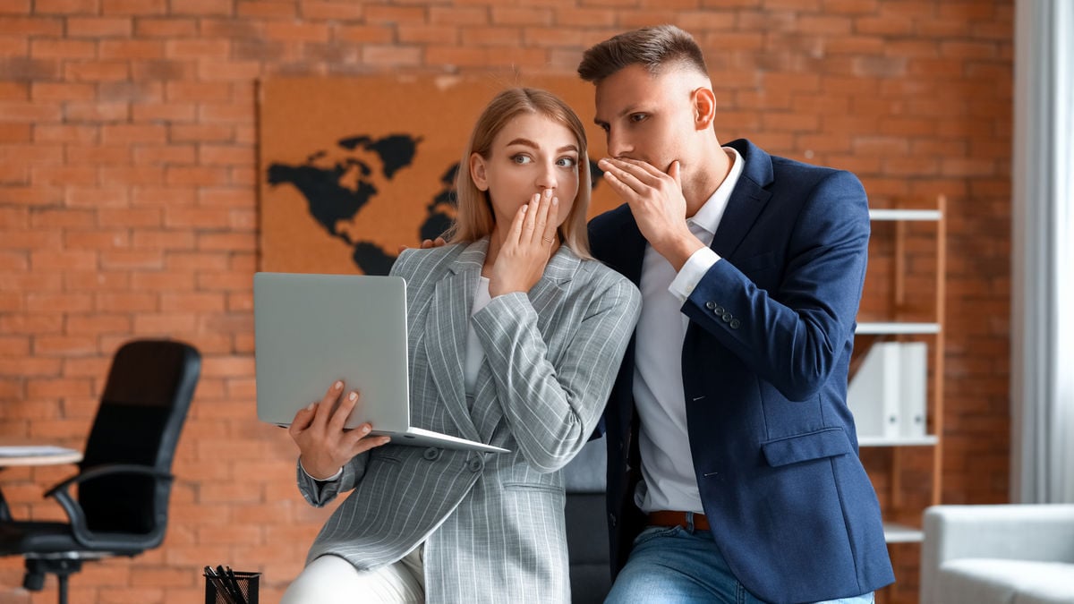 Two business colleagues sit together; one holds a laptop while both cover their mouths, whispering secrets. They are dressed in professional attire, with a brick wall and a world map in the background.