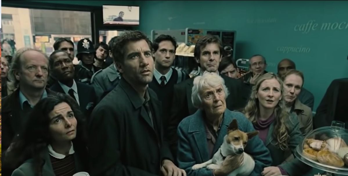 A group of people in a café stand closely together, looking up with serious and concerned expressions. An older woman in front holds a small dog. The teal wall behind displays words like "caffe mocha" and "cappuccino.