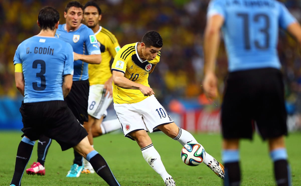 A soccer player in a yellow jersey, number 10, kicks the ball while surrounded by opponents in blue during a match, with focused players and a vibrant crowd in the background.