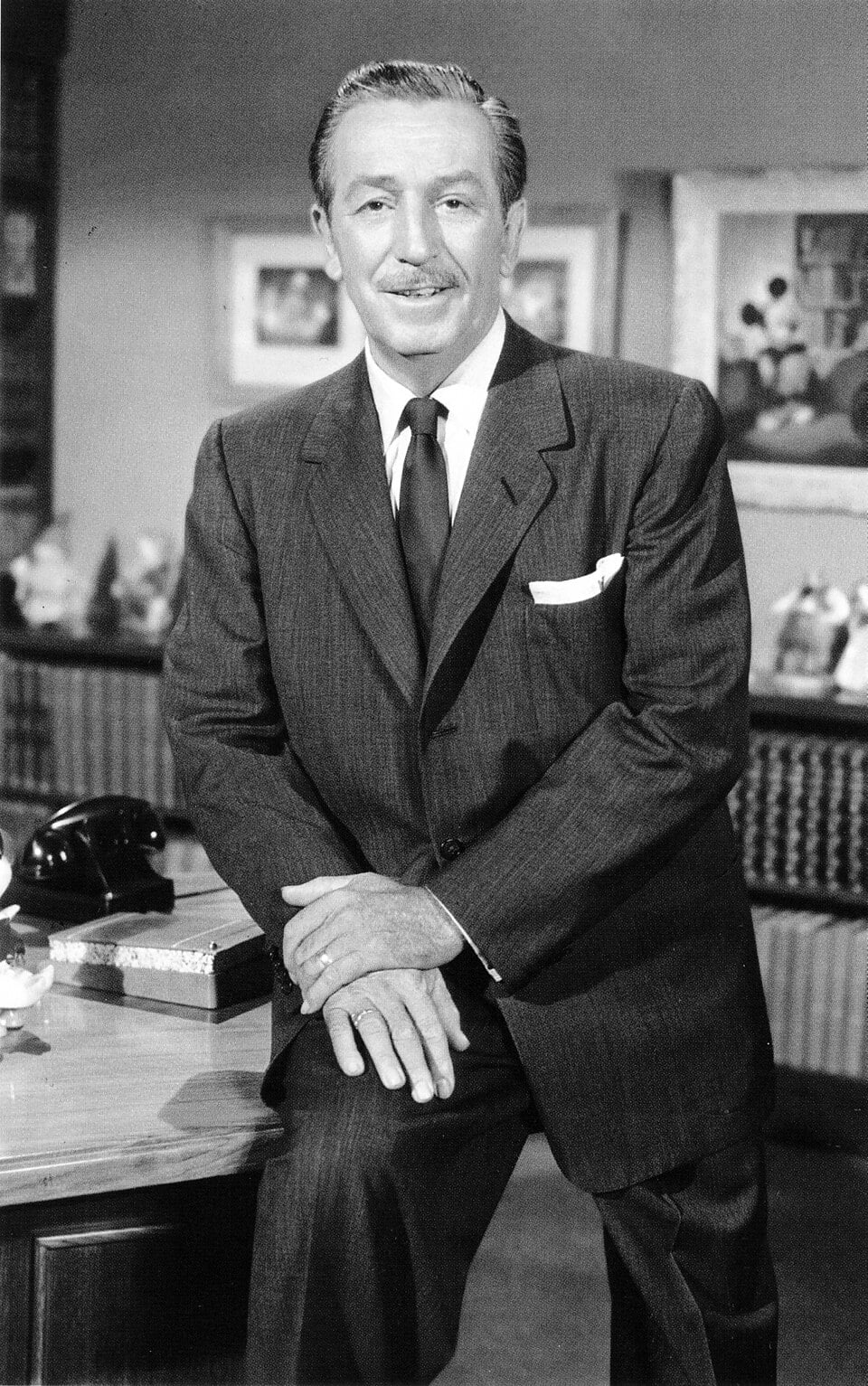 A man in a suit and tie with a mustache stands beside a desk, smiling at the camera. The room has framed pictures and shelves with small figurines in the background.