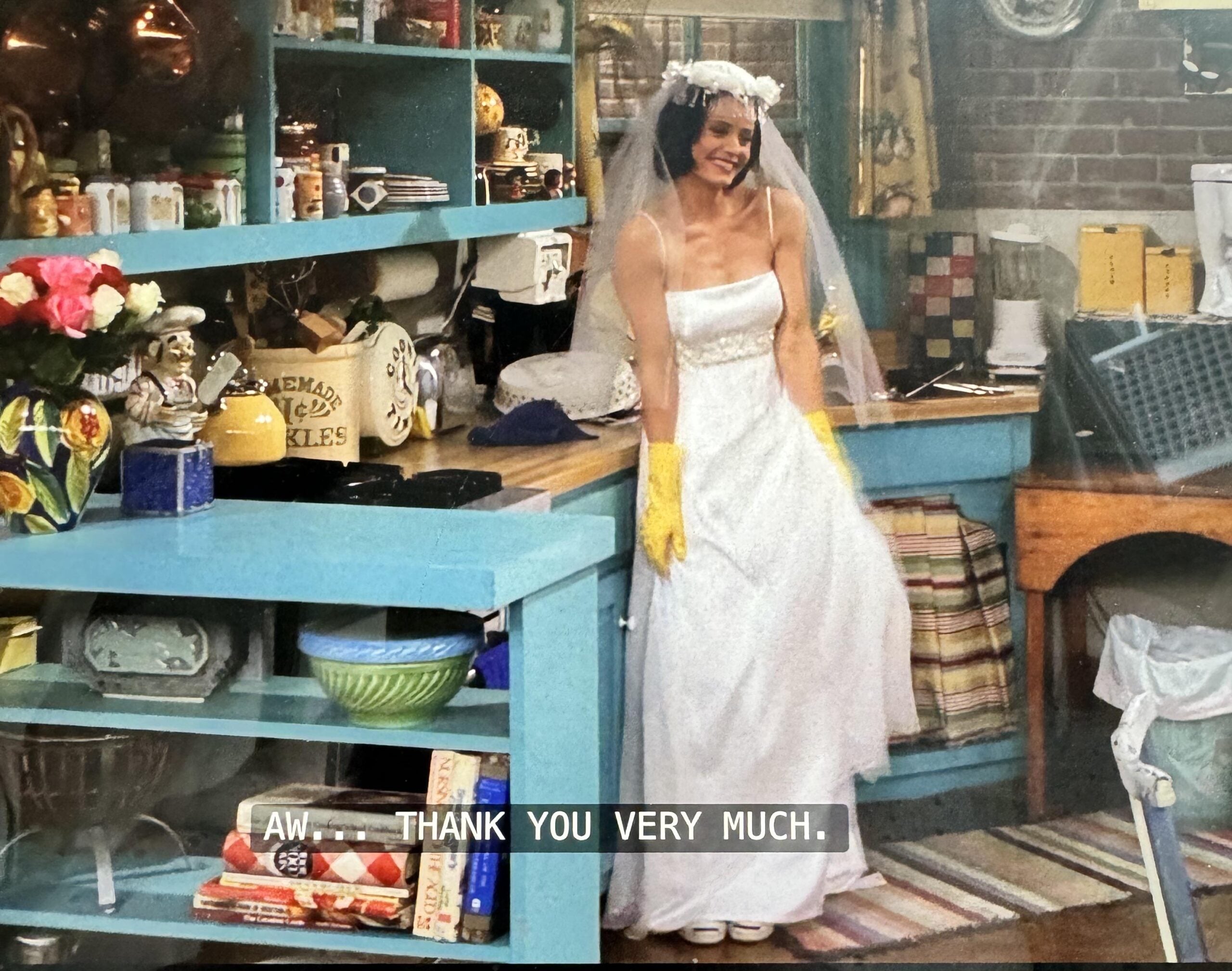 A woman in a wedding dress and veil, wearing yellow rubber gloves, stands smiling in a colorful kitchen. The kitchen shelves are cluttered with dishes and jars. Caption: "Aw... thank you very much.