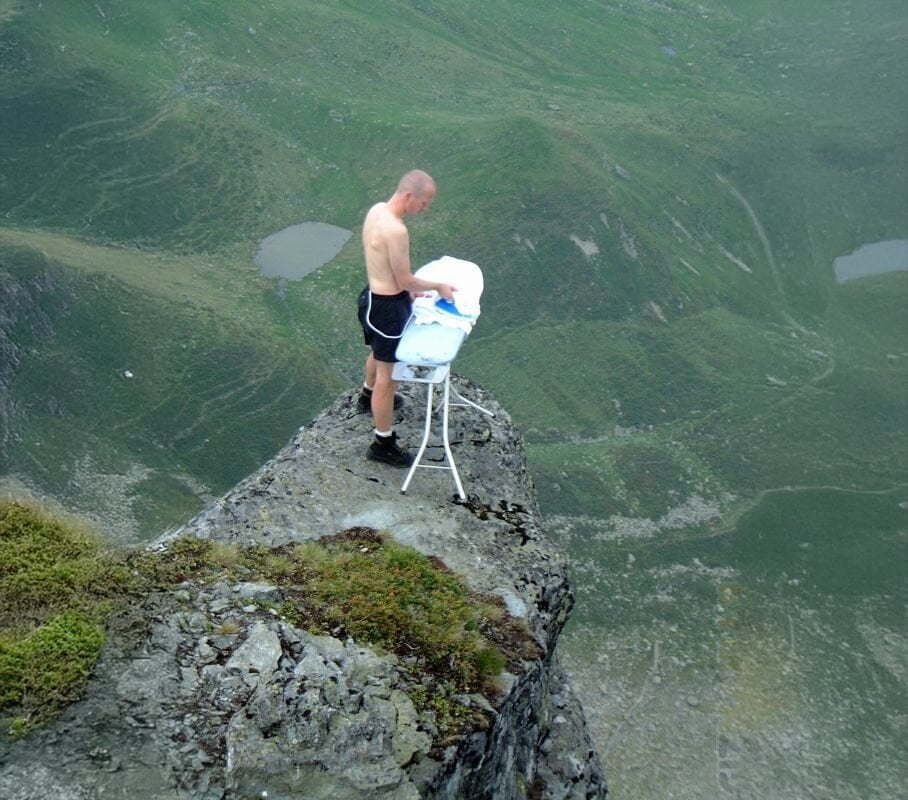 A shirtless man stands on the edge of a rocky cliff, wearing shorts, and irons clothes on an ironing board with a steep drop and green landscape below him.