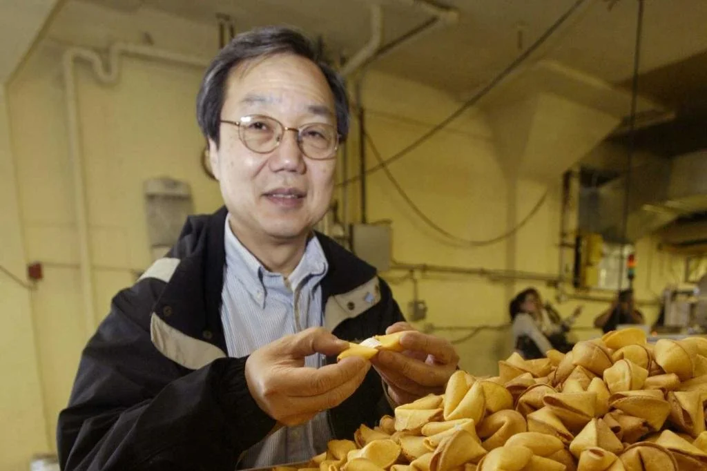 A person wearing glasses and a jacket stands next to a large pile of fortune cookies, holding one in their hand, inside what appears to be a factory or kitchen setting.
