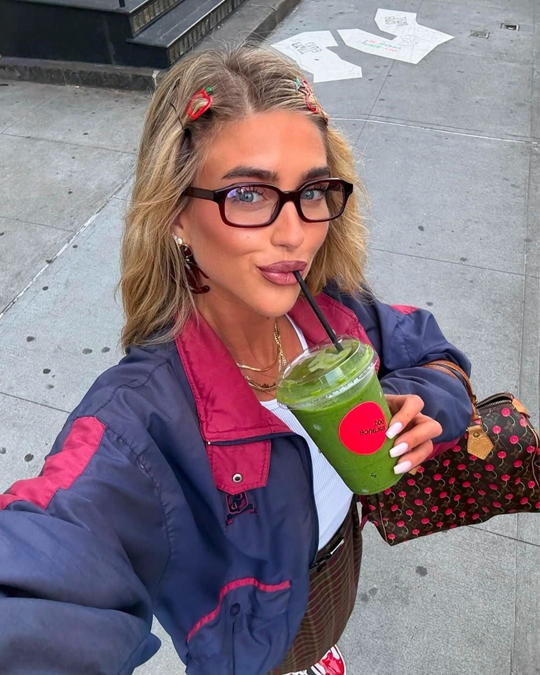 A woman with blonde hair, glasses, and hair clips takes a selfie outdoors while sipping a green smoothie through a straw. She wears a blue and burgundy jacket and carries a patterned bag. The sidewalk and street are visible behind her.