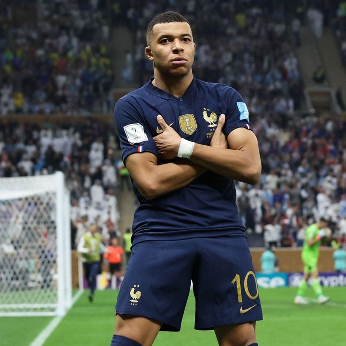 A soccer player in a navy blue France jersey with the number 10 stands in front of the goal, arms crossed over his chest, celebrating during a match, with a crowd in the background.