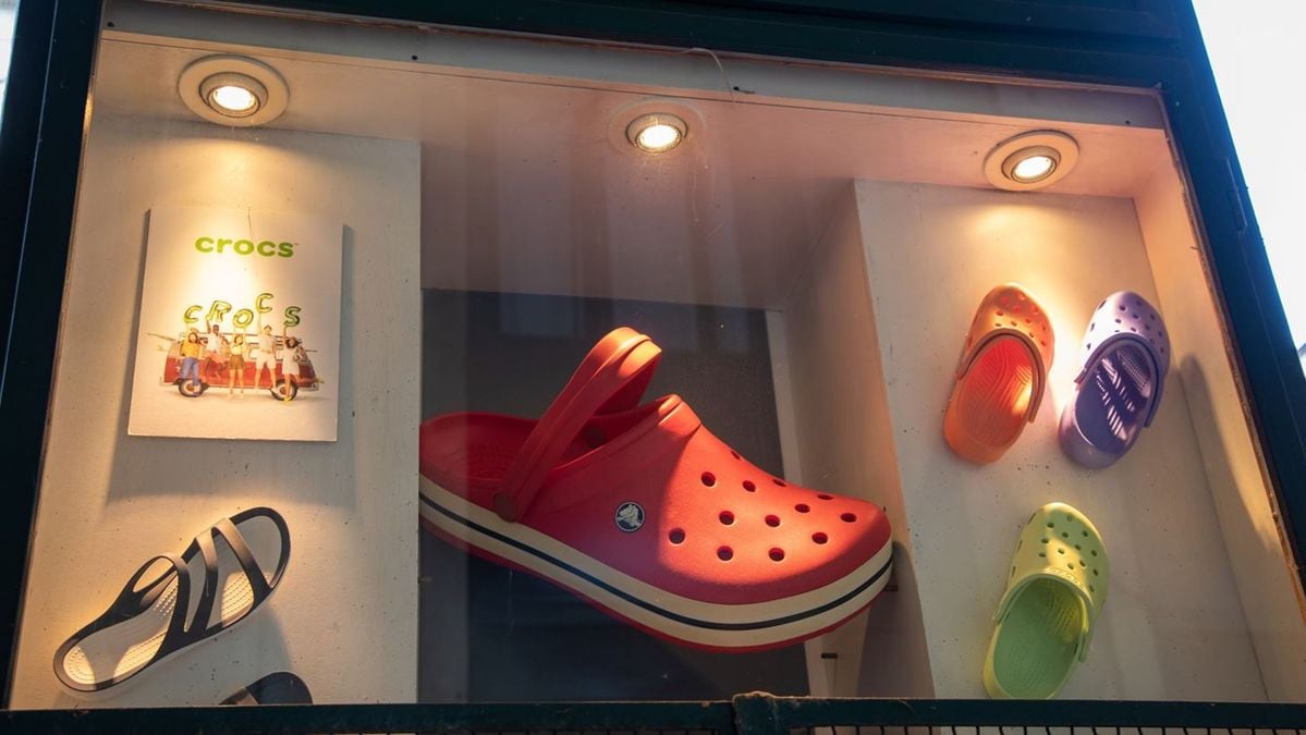 A store display featuring various Crocs sandals in different colors and sizes, with a large red Croc as the centerpiece, smaller Crocs on the sides, and a Crocs poster on the left wall, all under spotlights.