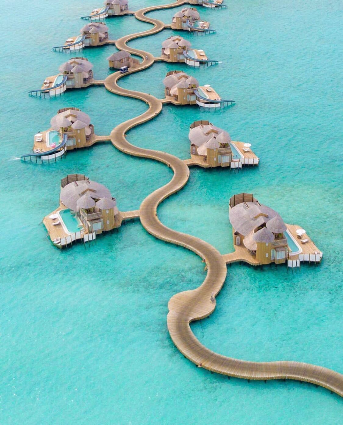 A winding wooden boardwalk connects several overwater villas, each with thatched roofs and private pools, above clear turquoise sea water. The scene is tranquil and tropical.