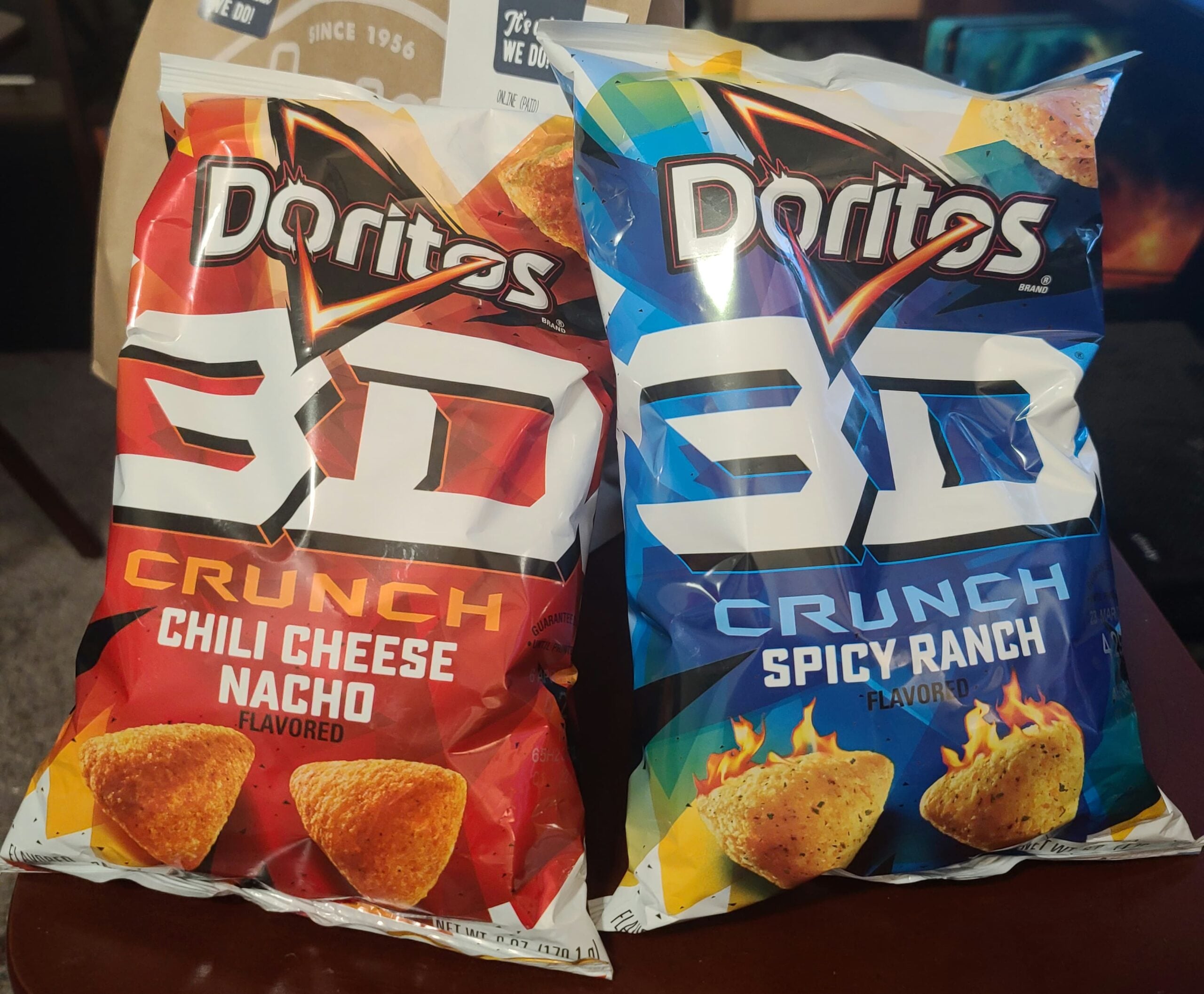 Two bags of Doritos 3D Crunch chips are side by side: one is Chili Cheese Nacho flavor in a red bag, and the other is Spicy Ranch flavor in a blue bag. Both bags feature images of the triangular chips.
