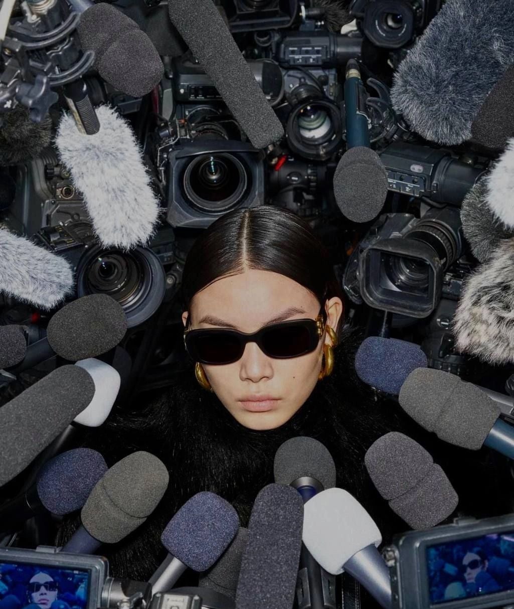 A person wearing sunglasses and a black coat is surrounded by numerous microphones and cameras, giving the impression of an intense media frenzy or press conference.