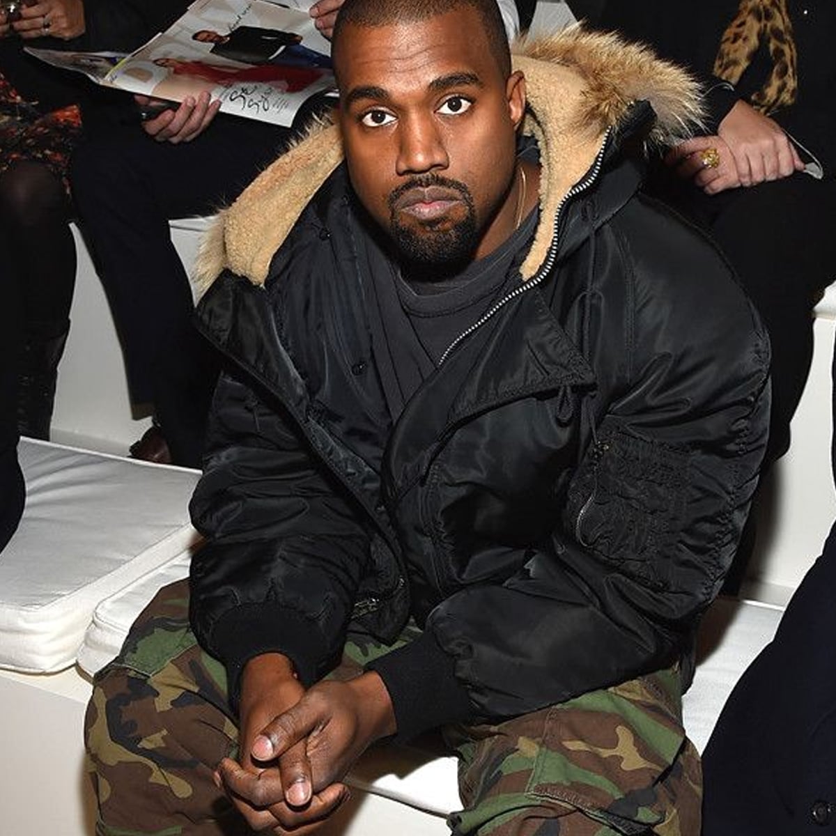 A man in a black jacket with a fur-lined hood and camouflage pants sits on a white bench, looking directly at the camera, with other people seated in the background.