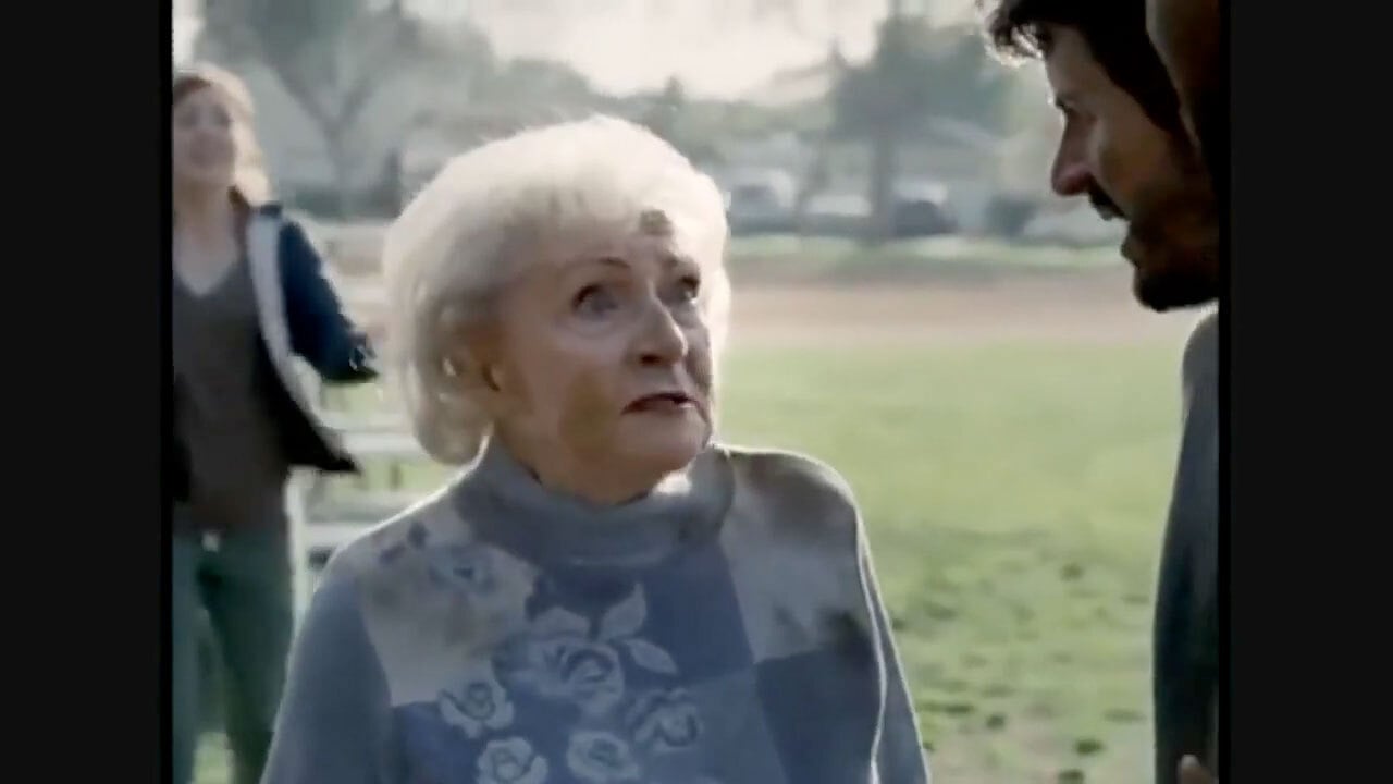 An elderly woman with white hair and a floral sweater looks up at a man outdoors in a park, with another person blurred in the background.