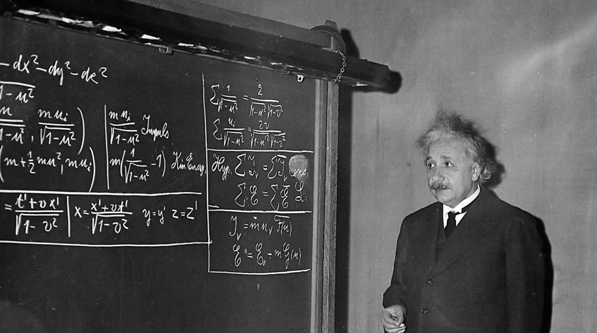 A man in a suit stands next to a chalkboard filled with complex mathematical equations, appearing to explain or teach the content.