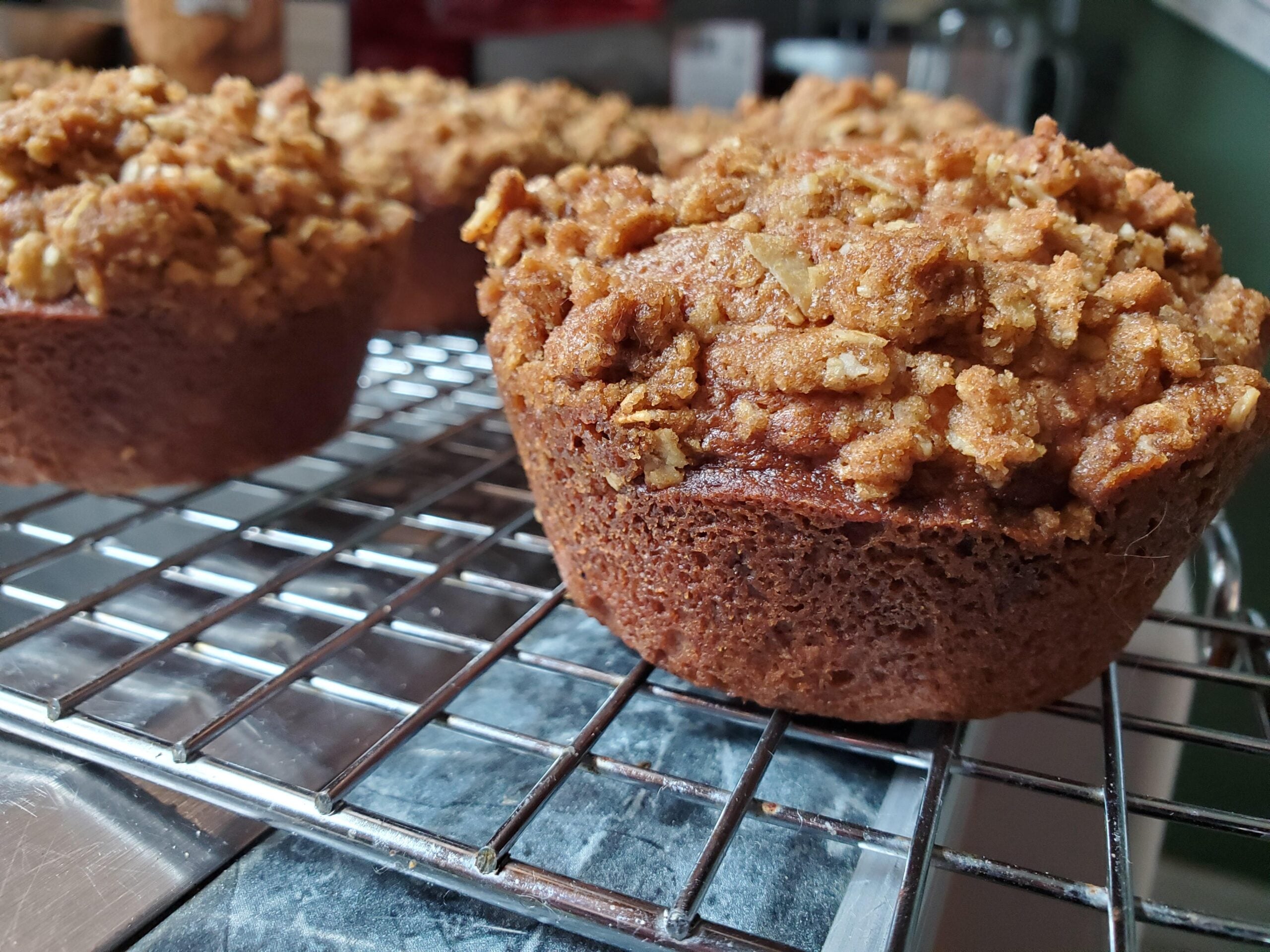 Close-up of a freshly baked muffin with a crumbly streusel topping, cooling on a metal wire rack. More muffins are visible in the background on the same rack.