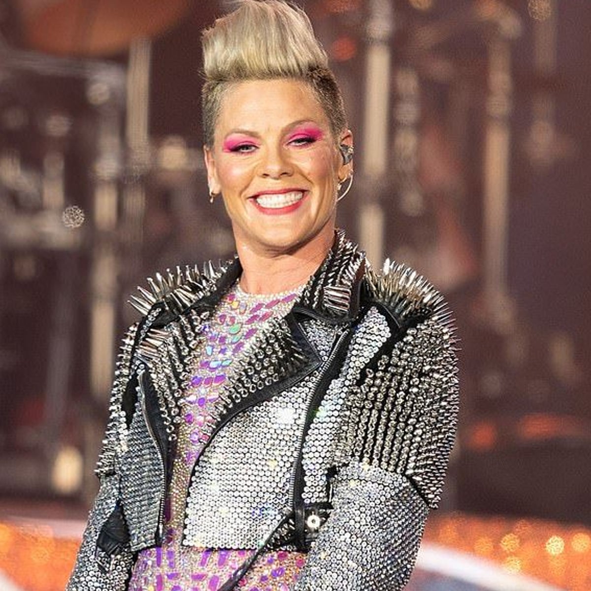 A smiling performer with short, spiked platinum hair wears a studded, sparkly jacket and bright pink eyeshadow, standing on stage with blurred lights and instruments in the background.