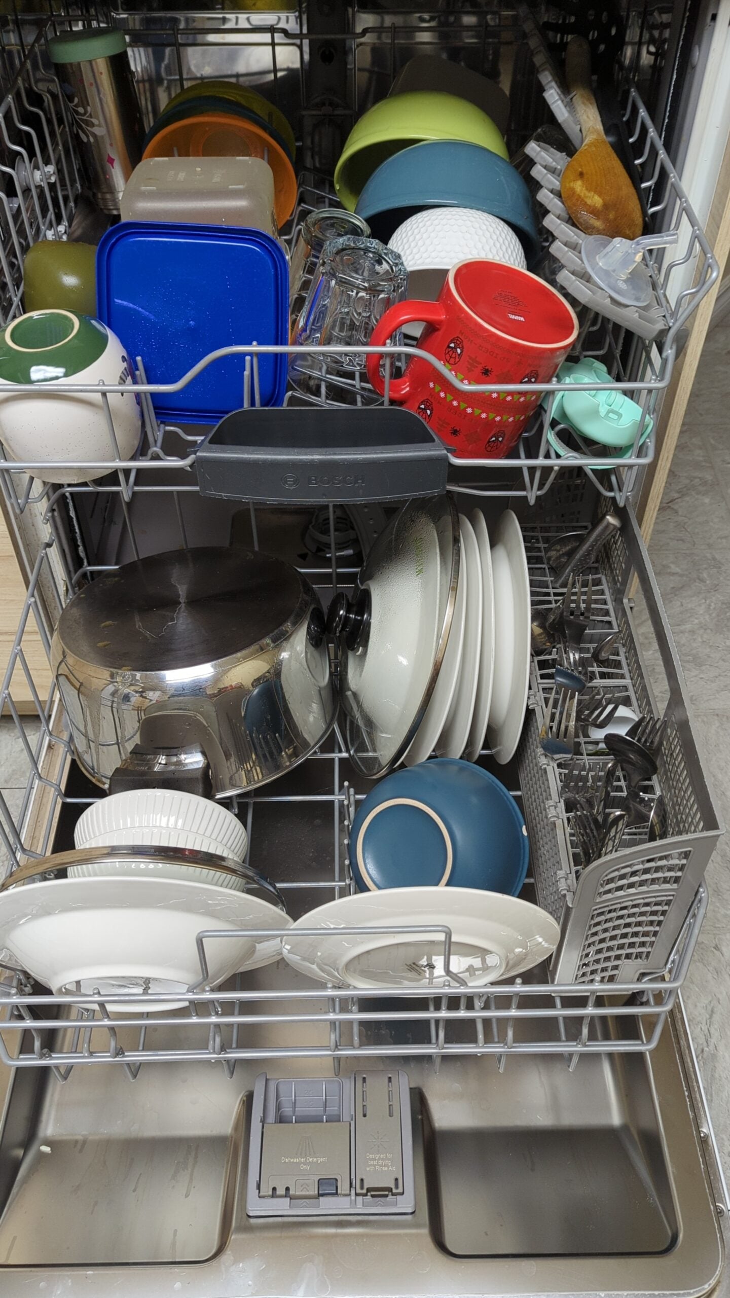 Open dishwasher filled with clean dishes, including plates, bowls, cups, mugs, lids, and various kitchenware items organized neatly on racks. The door is open, and the detergent compartment is visible.