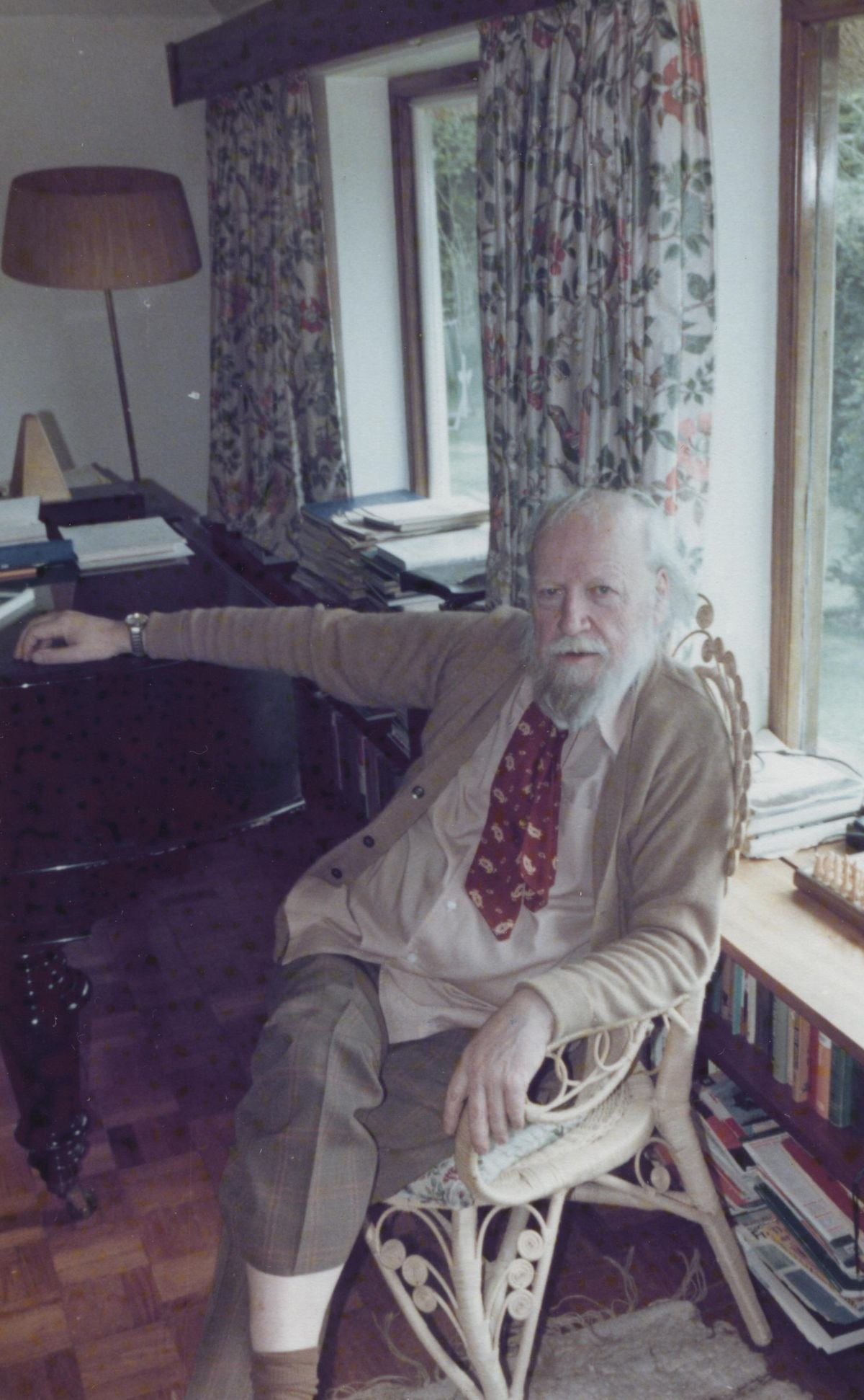 An elderly man with a white beard sits in a wicker chair by a window, wearing a tan cardigan, red patterned tie, and brown pants. He is in a cozy room with books, a desk, and floral curtains.