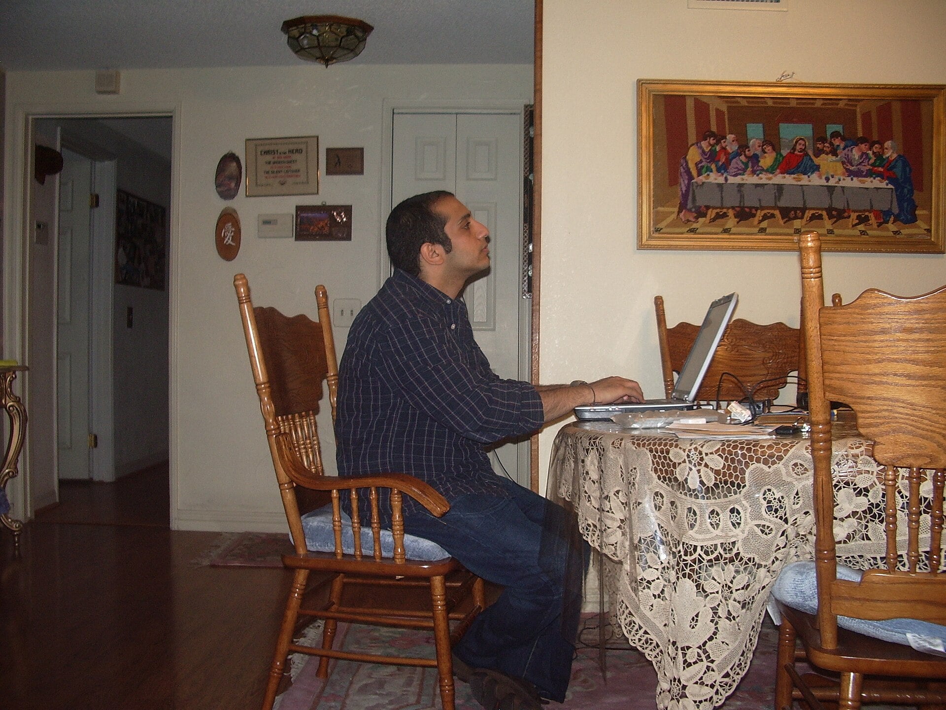 A man sits at a dining table using a laptop in a cozy room with wooden chairs, a lace tablecloth, and artwork on the wall, including a framed picture of The Last Supper.