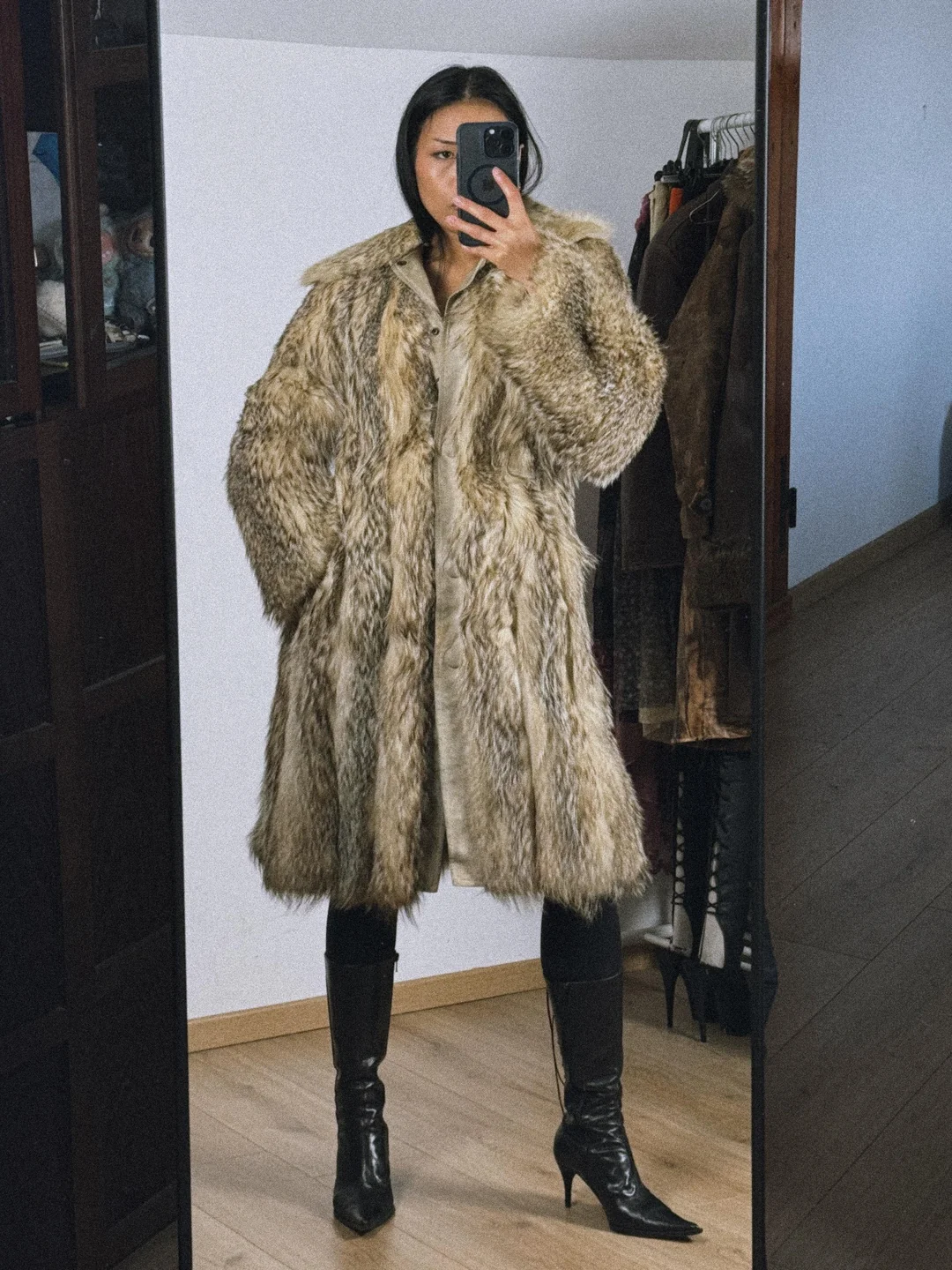 A person stands in front of a mirror taking a selfie, wearing a long, shaggy fur coat, black heeled boots, and black pants. Clothing and jackets are visible in the background.