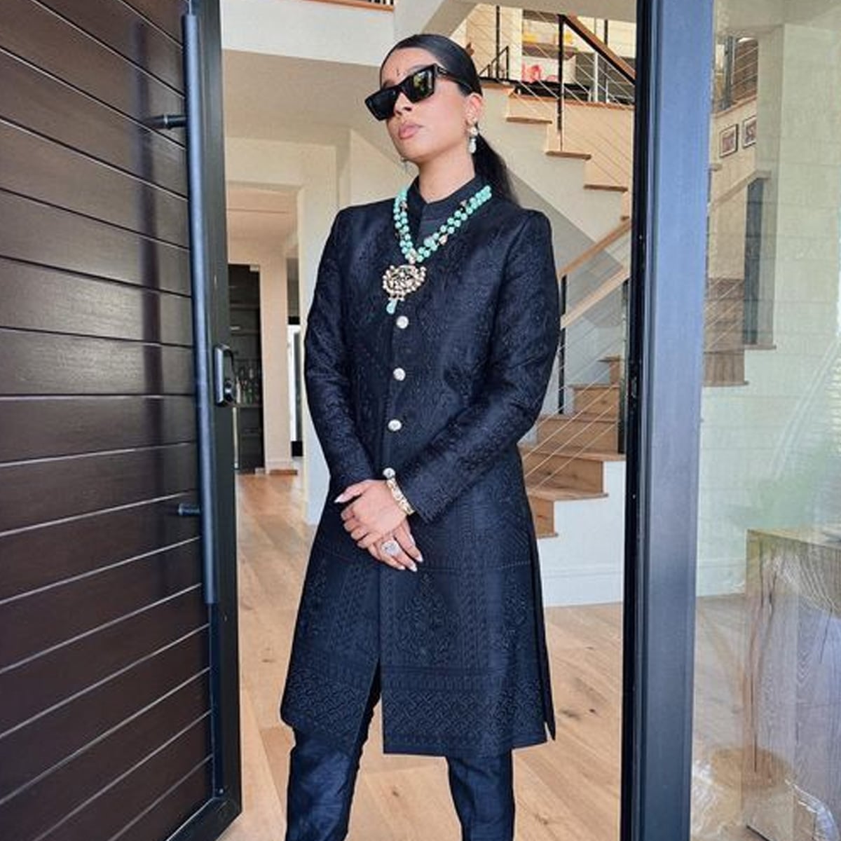 A woman wearing a long black embroidered coat, black pants, statement necklace, and black sunglasses stands confidently in a modern home entrance with a staircase in the background.