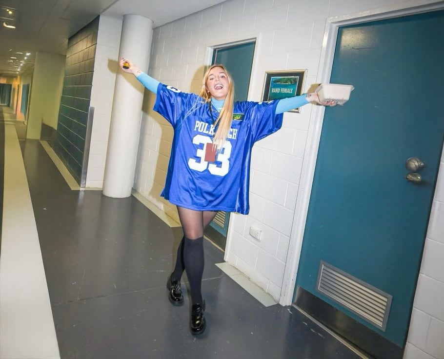 A woman in a blue oversized sports jersey with "33" on it stands in a corridor, smiling with arms outstretched, holding food. She wears black tights and shoes, and appears joyful.