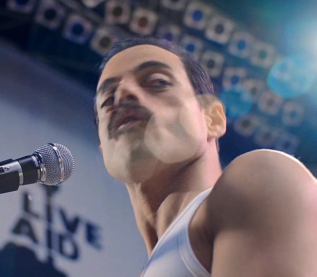 A person with a mustache wearing a white tank top stands at a microphone on stage, with "LIVE AID" visible in the blurred background and bright stage lights overhead.