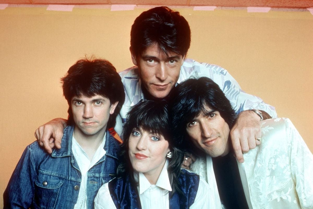 Four people pose together in front of a yellow background, with two standing in back and two sitting in front. All have dark hair and are wearing casual or semi-formal clothing, smiling or looking at the camera.
