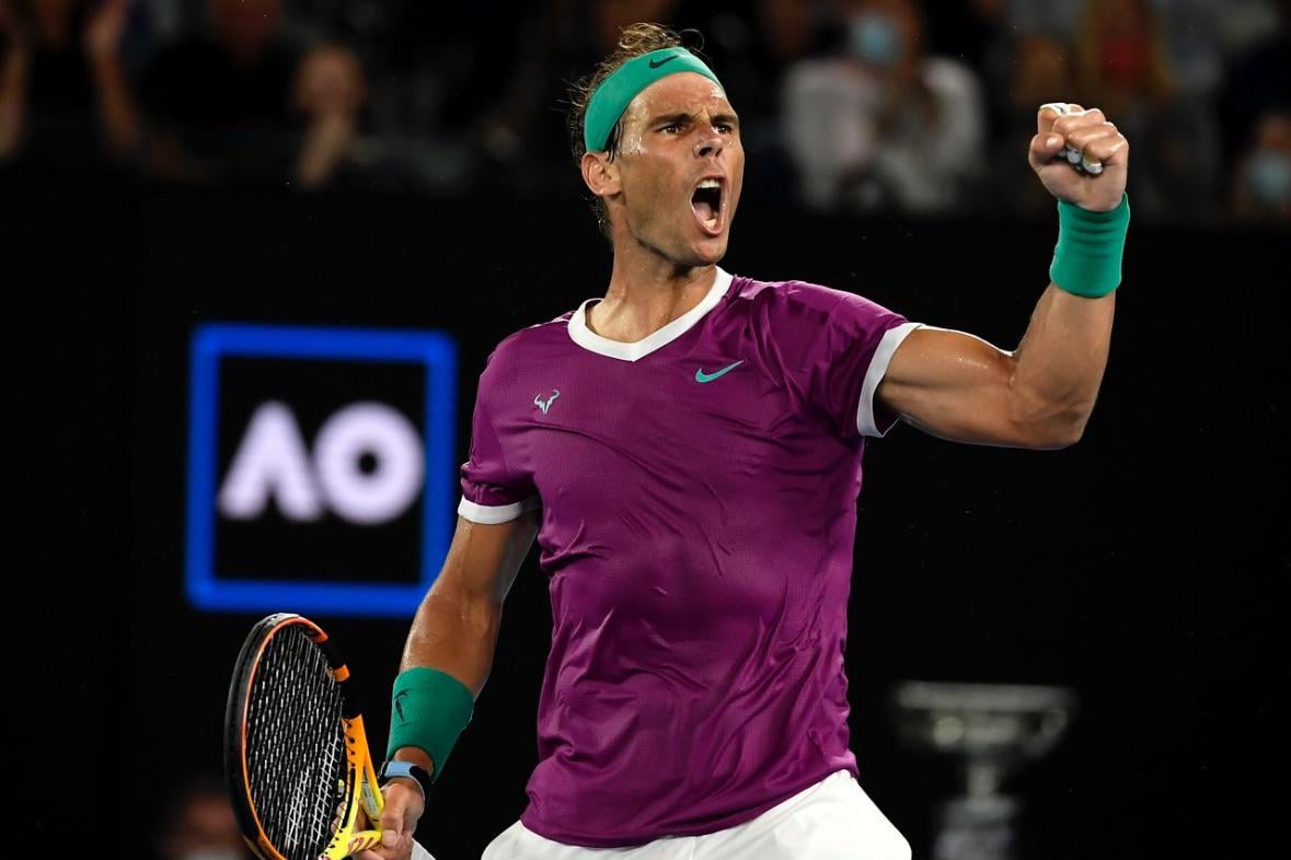 A male tennis player in a purple shirt pumps his fist and shouts in celebration on a tennis court, holding a racket. The background shows a blurred crowd and an illuminated "AO" sign.