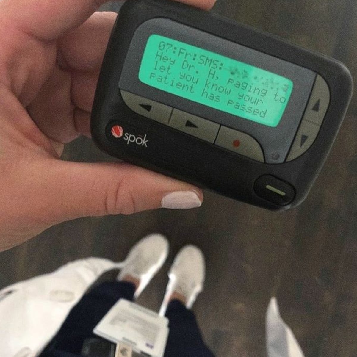 A hand holds a pager displaying a message: "Hey Dr. H, paging to let you know your patient has passed." The person is wearing white shoes and a lab coat, visible from a top-down view.