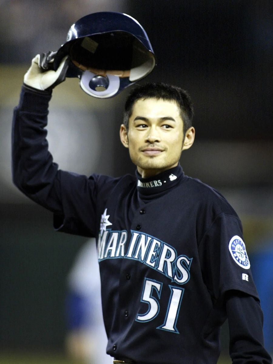 A baseball player in a Seattle Mariners uniform holds his helmet above his head and smiles, wearing jersey number 51.