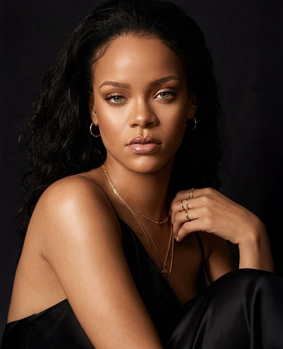 A woman with long, wavy dark hair poses against a black background, wearing a black satin top, gold necklaces, hoop earrings, and several gold rings, with a neutral facial expression.