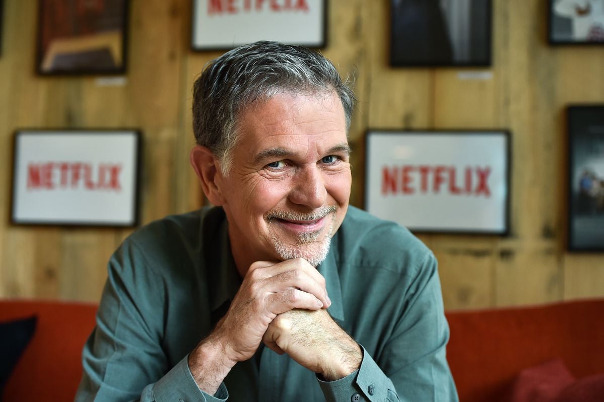 A smiling man with gray hair and a goatee rests his chin on his hands. He sits on an orange couch with Netflix logos displayed on framed screens behind him on a wooden wall.