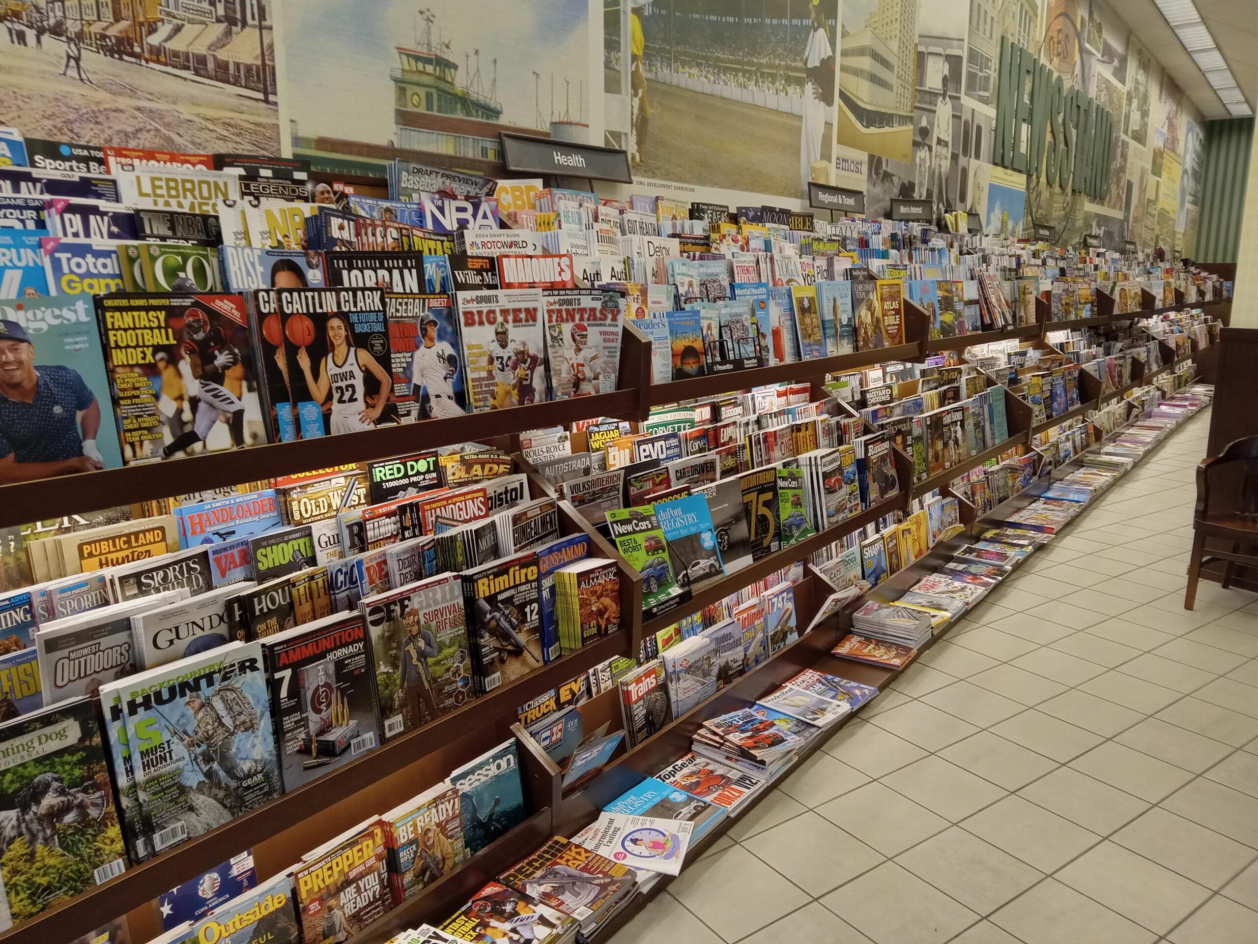 A large magazine rack in a bookstore displays a wide variety of magazines on shelves, featuring topics like sports, health, travel, and current events, with colorful covers filling the long aisle.