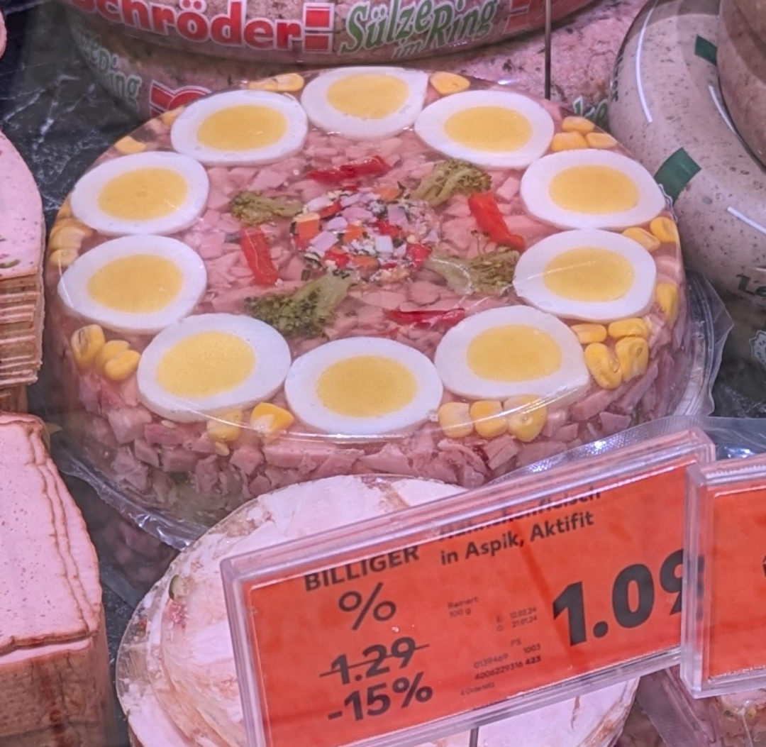 A round meat and vegetable aspic topped with sliced boiled eggs, corn, and vegetables is displayed in a deli case next to slices of luncheon meat. An orange price tag in German indicates a discount price of 1.08 euros.