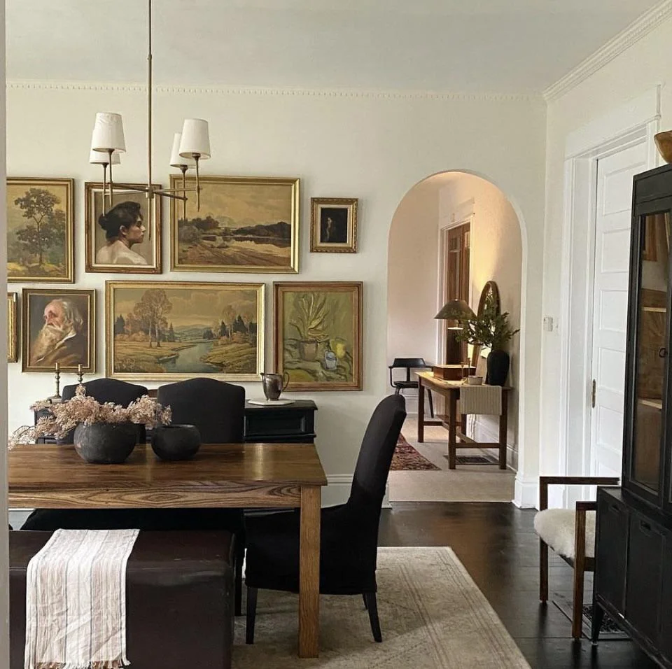 A dining room with a wooden table, dark chairs, and a light runner. The wall displays multiple framed paintings, including portraits and landscapes. An arched doorway leads to a hallway with a side table and plants.