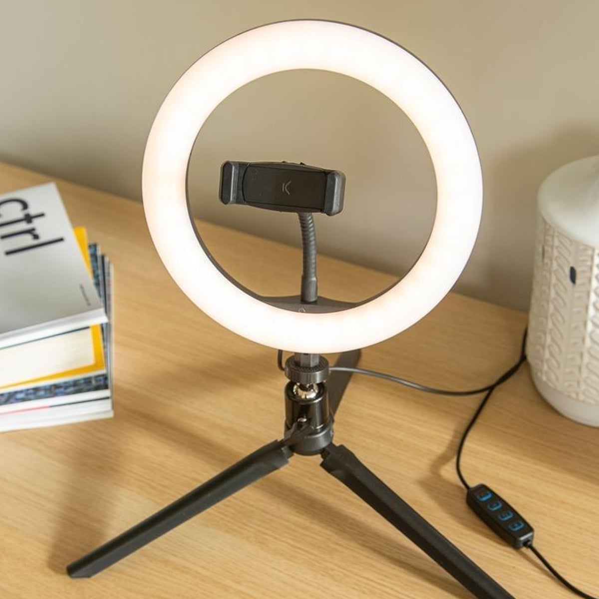 A lit ring light with a phone holder in the center is set up on a wooden desk. A small tripod supports the light, and a remote control is attached to the power cord. Nearby are stacked books and a white lamp.