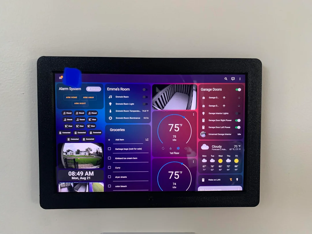 A smart home control panel mounted on a wall displays various controls and information, including alarm system status, room temperatures, garage door statuses, a grocery list, current time, date, and weather.