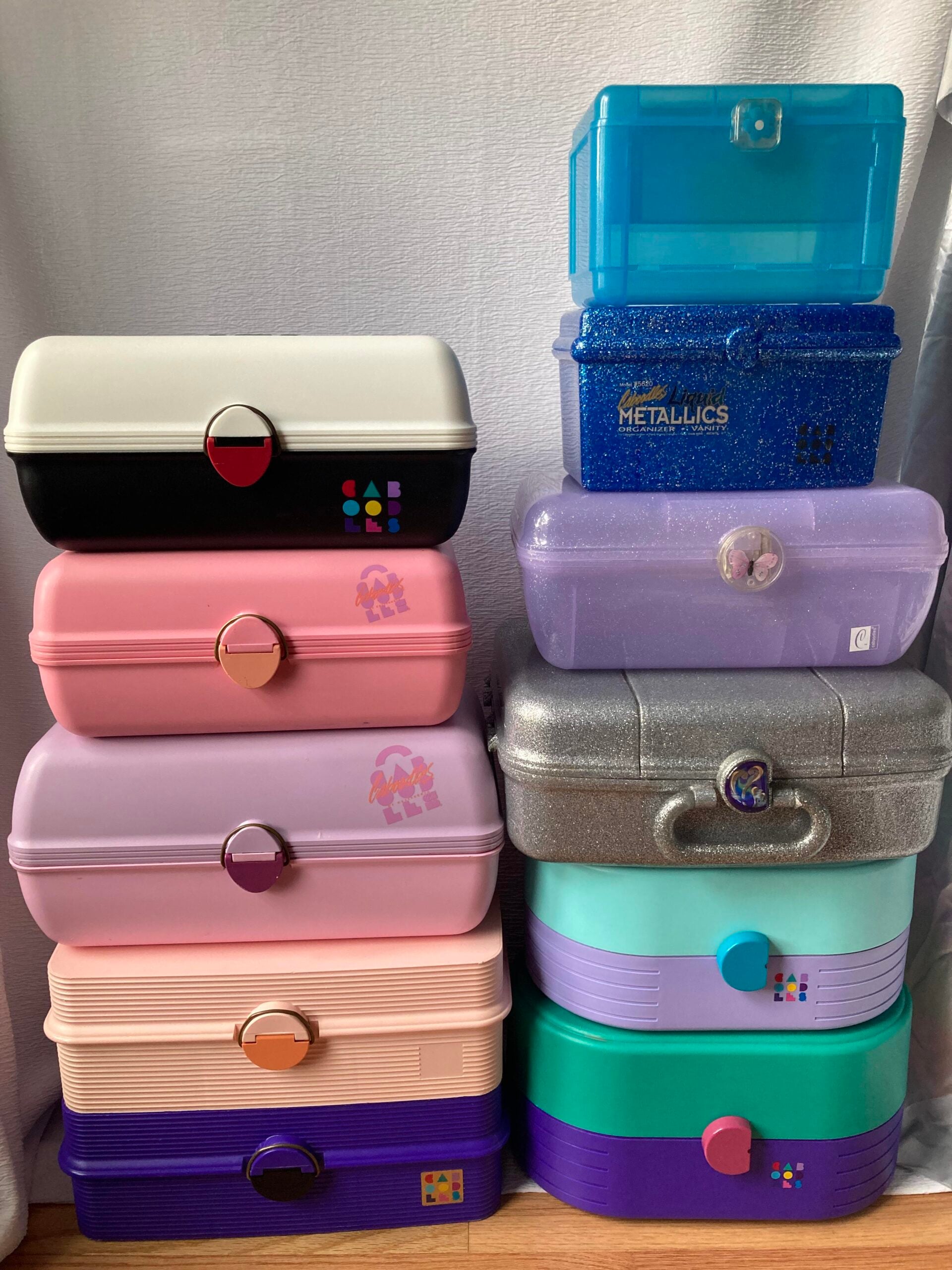 A colorful stack of Caboodles makeup organizers in various shapes and sizes sits against a white backdrop, featuring pink, purple, teal, blue, and glittery cases with different clasps and logos.