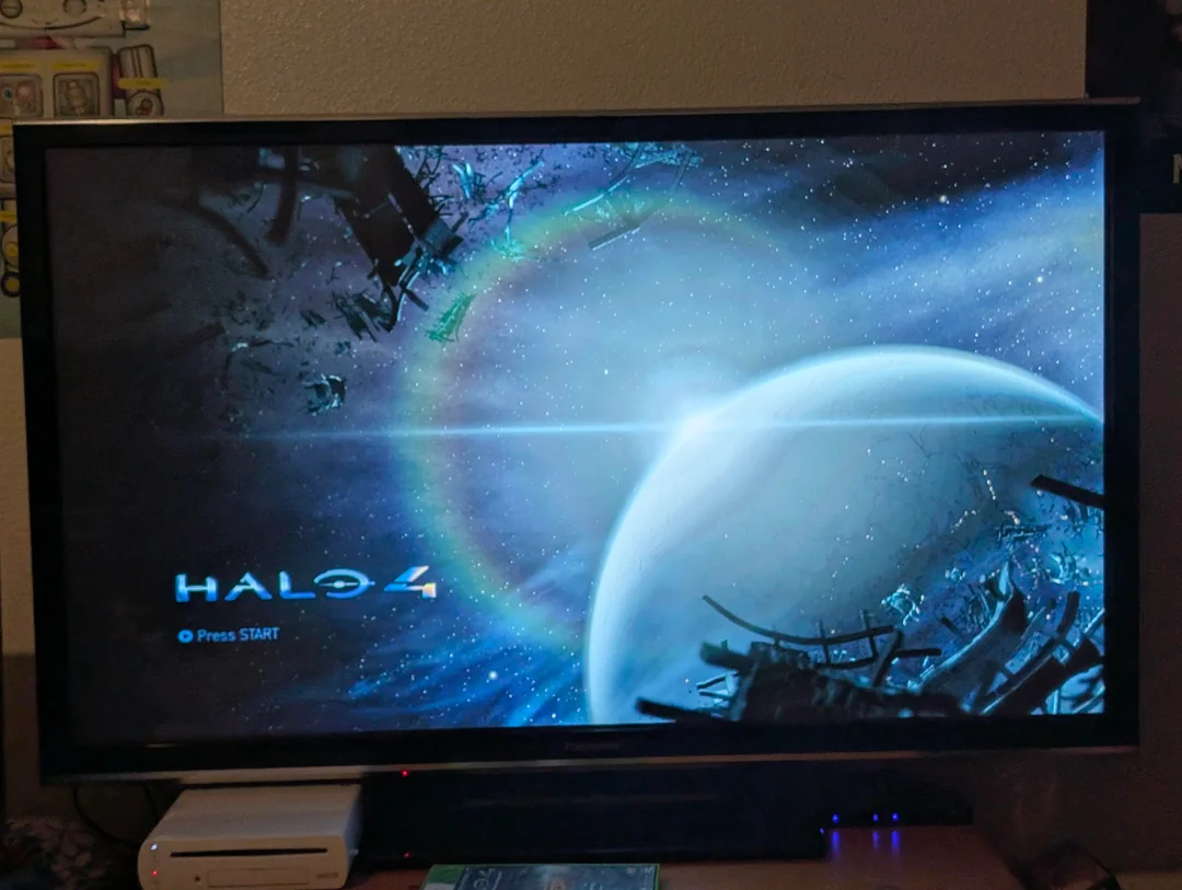 A television screen displays the Halo 4 start menu, showing a blue planet with surrounding space debris and a glowing ring in space. The "Press START" prompt is visible in the lower left corner.