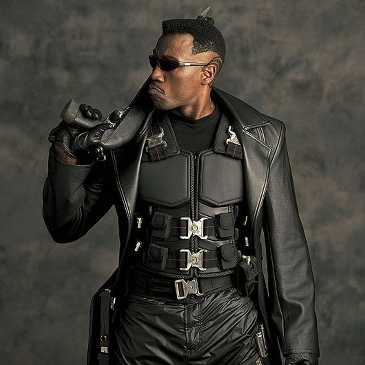 A man in a black tactical outfit, leather coat, and sunglasses holds a sword over his shoulder, looking serious against a dark, textured background.