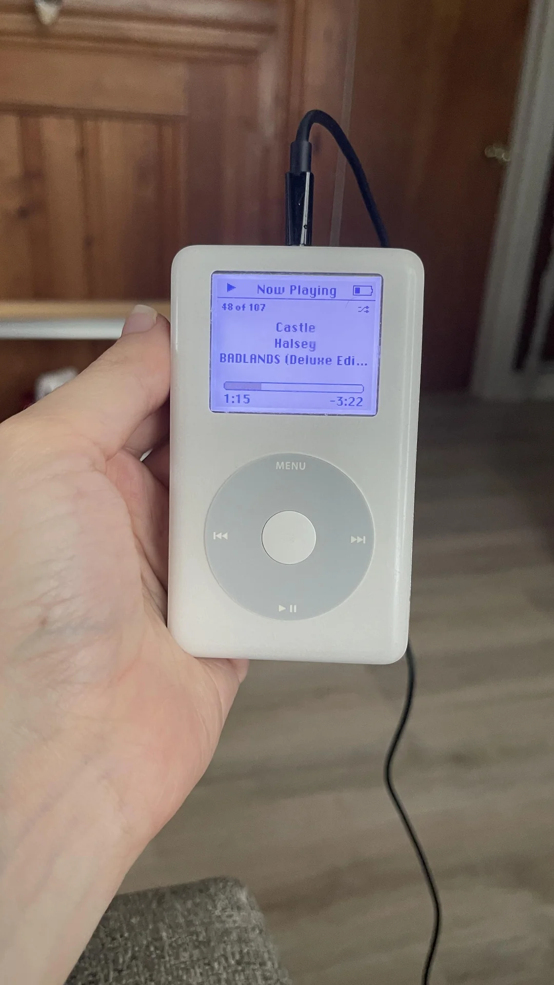 A hand holds a white iPod Classic displaying the song "Castle" by Halsey from the album "BADLANDS (Deluxe Edition)" on its screen. The device is connected to headphones.