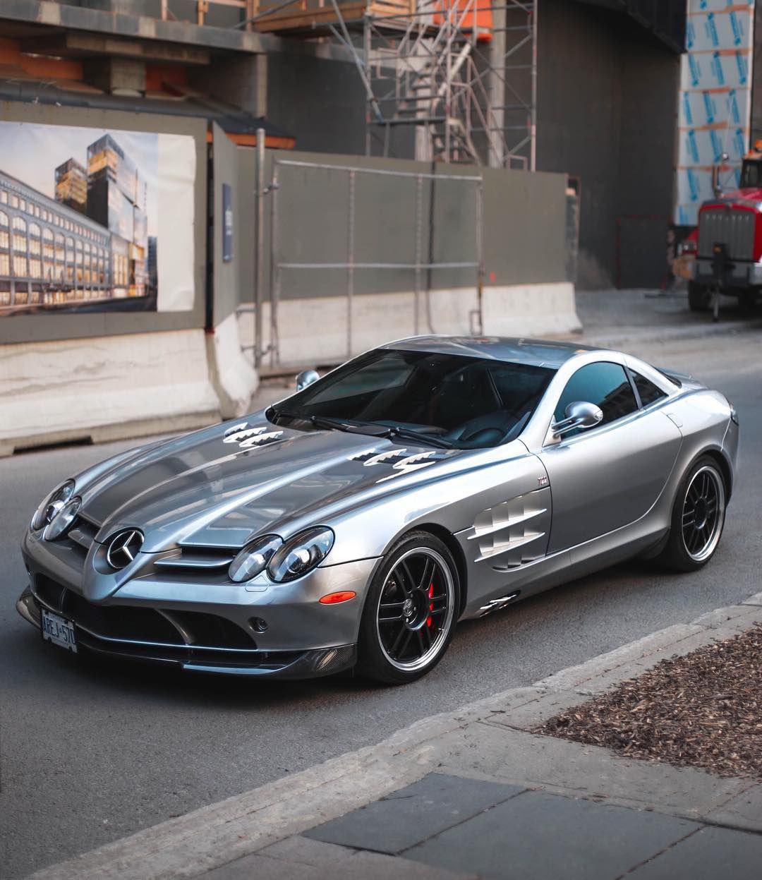 A silver Mercedes-Benz SLR McLaren sports car is parked on a city street near a construction site, with scaffolding and barriers visible in the background.