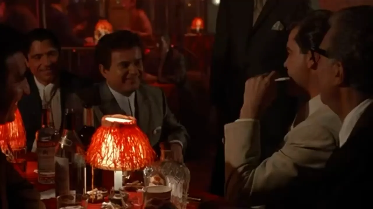 Four men in suits sit around a dimly lit table in a restaurant or club, with a red-shaded lamp, drinks, and cigarettes on the table. One man is smiling, and the scene has a warm, intimate atmosphere.
