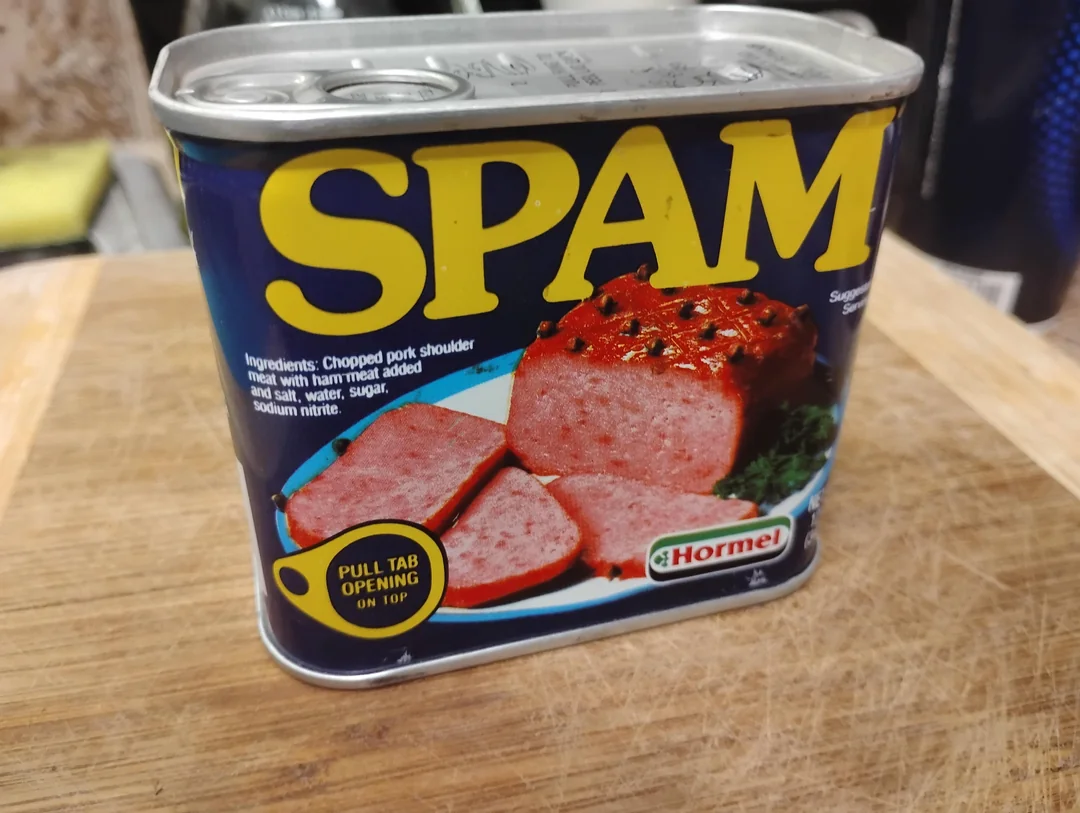 A can of SPAM with a pull tab sits on a wooden surface. The label shows sliced SPAM on a plate with garnish. The ingredients and Hormel logo are visible.